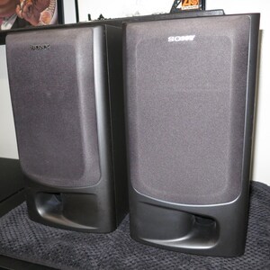 May include: A pair of black Sony bookshelf speakers featuring a rectangular design with a fabric grille covering the upper section and a bass reflex port located at the bottom front of each unit. The Sony logo is printed in white at the top.
