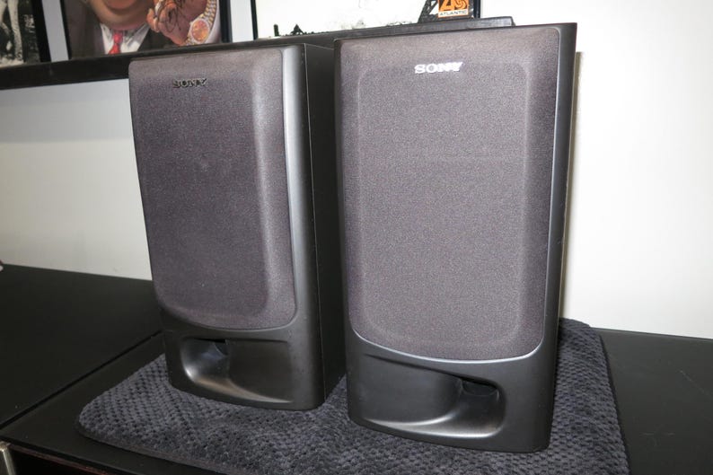 May include: A pair of black Sony bookshelf speakers featuring a rectangular design with a fabric grille covering the upper section and a bass reflex port located at the bottom front of each unit. The Sony logo is printed in white at the top.