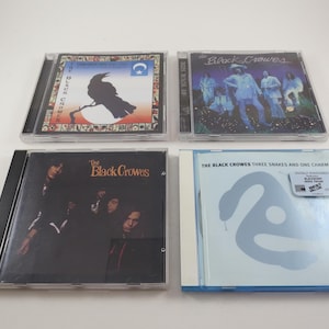May include: Four compact discs by the band The Black Crowes. The first disc has a black crow on a branch with a blue and white background. The second disc has a photo of the band members on a blue and purple background. The third disc has a photo of the band members on a black background. The fourth disc has a blue and white swirl design with the text "The Black Crowes Three Snakes and One Charm".