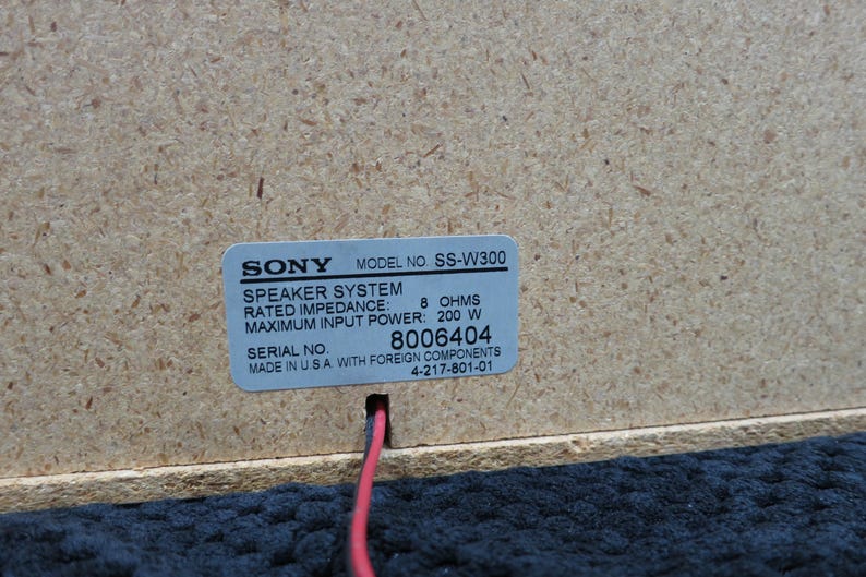 May include: Close-up of a speaker system component. A label with the "SONY" logo and model number "SS-W300" is affixed to a light brown wooden surface. The label includes technical specifications and a serial number.