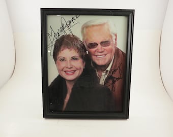 May include: A black framed photo of a man and woman. The man is wearing a brown jacket and sunglasses. The woman is wearing a black jacket. Both are smiling and have signed their names on the photo.