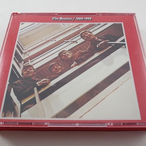 May include: A red CD case featuring a photograph of The Beatles. The image shows the band members looking down from a balcony. The text "The Beatles / 1962-1966" is printed at the top of the case.