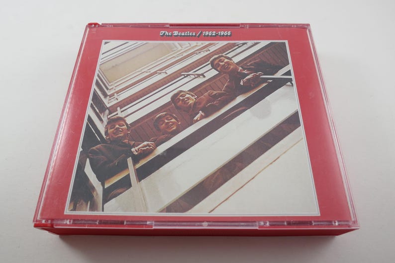 May include: A red CD case featuring a photograph of The Beatles. The image shows the band members looking down from a balcony. The text "The Beatles / 1962-1966" is printed at the top of the case.