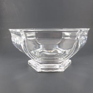 May include: A clear glass bowl featuring a multi-faceted, octagonal design with a wide, open top and a sturdy, raised base. The transparent glass construction displays smooth, curved sides that transition into sharp, geometric angles at the bottom.