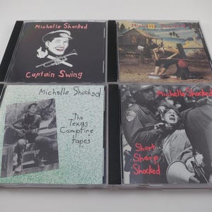 May include: Four Michelle Shocked music CDs. The album covers feature text and images, including the titles "Captain Swing", "Arkansas Traveler", "The Texas Campfire Tapes", and "Short Sharp Shocked". The covers are in clear plastic cases.