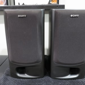 May include: A pair of black Sony bookshelf speakers with rectangular fabric grilles and a bottom-facing bass reflex port. Each speaker features the Sony brand name in silver lettering at the top center of the front panel, with a solid black frame.