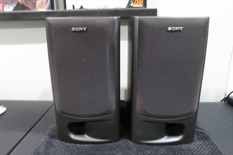 May include: A pair of black Sony bookshelf speakers with rectangular fabric grilles and a bottom-facing bass reflex port. Each speaker features the Sony brand name in silver lettering at the top center of the front panel, with a solid black frame.