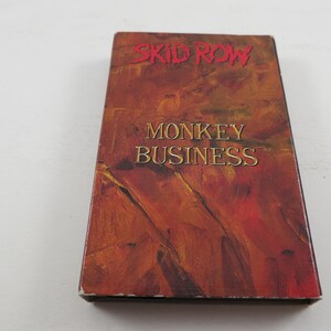 May include: A VHS cassette tape with an abstract design in shades of red, orange and yellow. The text "SKID ROW" and "MONKEY BUSINESS" is printed on the front of the tape.