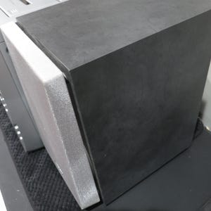 May include: A black rectangular speaker with a gray foam pad attached to its side. The speaker is positioned next to a silver component. The speaker is on a black surface.