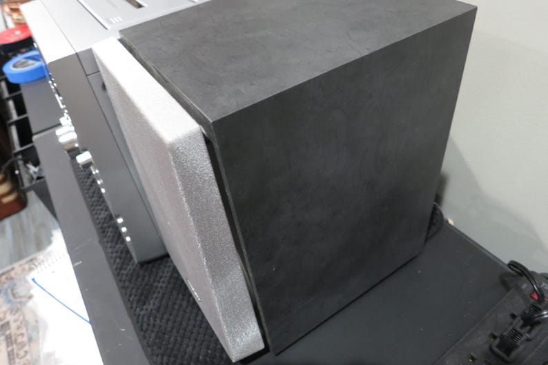 May include: A black rectangular speaker with a gray foam pad attached to its side. The speaker is positioned next to a silver component. The speaker is on a black surface.