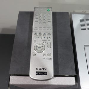 May include: Silver Sony AV System remote control with numerous buttons, including a green power button. The remote is labeled "RM-SS300" and rests on a dark gray surface, next to a silver electronic device.