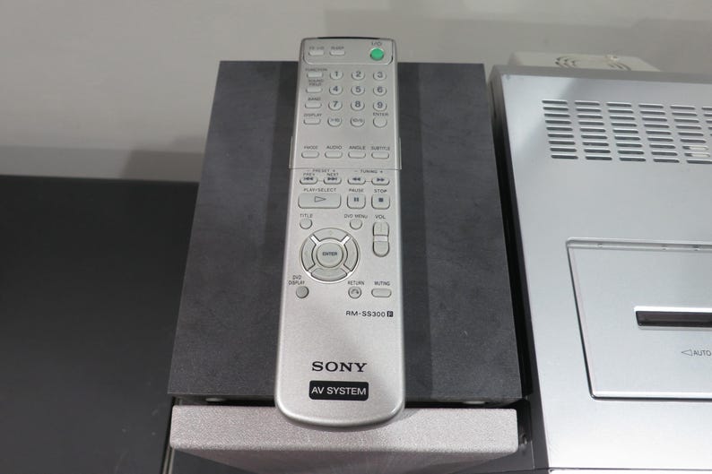 May include: Silver Sony AV System remote control with numerous buttons, including a green power button. The remote is labeled "RM-SS300" and rests on a dark gray surface, next to a silver electronic device.