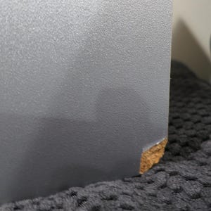 以下が含まれることがあります： 質感のある仕上げが施されたグレーの平らな物体のクローズアップ。物体の下隅には、グレーの外層が欠けたり摩耗したりして、内部の明るい茶色の粒状の素材が露出している小さな箇所が見られます。