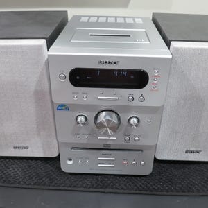 May include: A silver Sony stereo system with two gray speakers and a central unit. The unit has a CD player, digital display, and various control buttons. The speakers are rectangular and the system sits on a black surface.