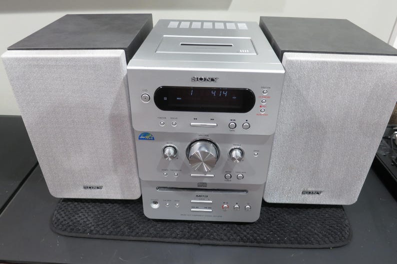 May include: A silver Sony stereo system with two gray speakers and a central unit. The unit has a CD player, digital display, and various control buttons. The speakers are rectangular and the system sits on a black surface.