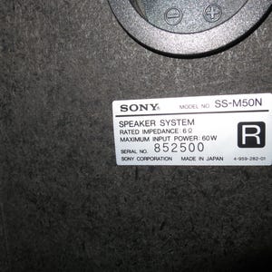 May include: A black Sony SS-M50N speaker system label with white text on a dark particle board surface. The label specifies a rated impedance of 6 ohms, a maximum input power of 60W, serial number 852500, and the text Made in Japan.