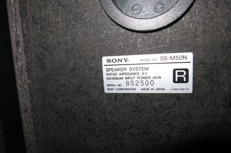 May include: A black Sony SS-M50N speaker system label with white text on a dark particle board surface. The label specifies a rated impedance of 6 ohms, a maximum input power of 60W, serial number 852500, and the text Made in Japan.