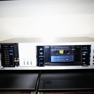 May include: A silver JVC stereo cassette deck featuring a front-loading tape compartment with a visible cassette tape labeled ThinkAdotOmY. The unit includes a black peak level display, multiple control buttons in black, blue, and red, and a tape counter.