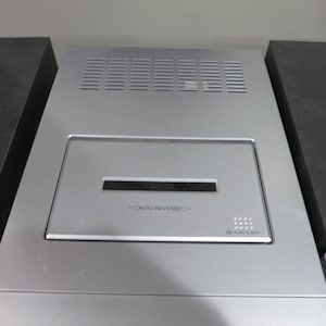 May include: Silver-colored electronic device with a rectangular design. Features a central panel with a slot and the text "AUTO REVERSE." The top has ventilation slits. Flanked by two dark gray, rectangular blocks.