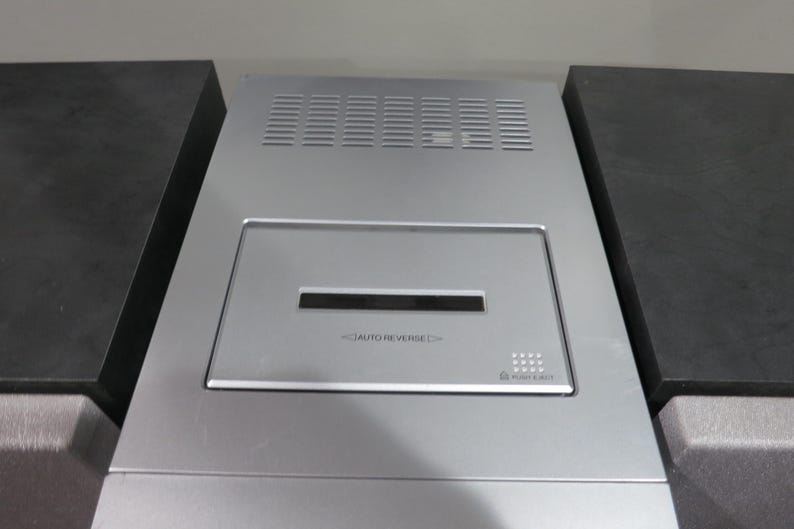 May include: Silver-colored electronic device with a rectangular design. Features a central panel with a slot and the text "AUTO REVERSE." The top has ventilation slits. Flanked by two dark gray, rectangular blocks.