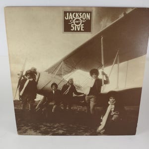 May include: Vintage album cover featuring the Jackson 5 posing in front of a biplane. The sepia-toned image shows the group in aviator attire, with the album title "Jackson 5" in a rectangular box at the top.