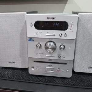 May include: A silver Sony stereo system with two speakers. The stereo has a digital display and various buttons and knobs. The speakers and stereo are on a black surface.