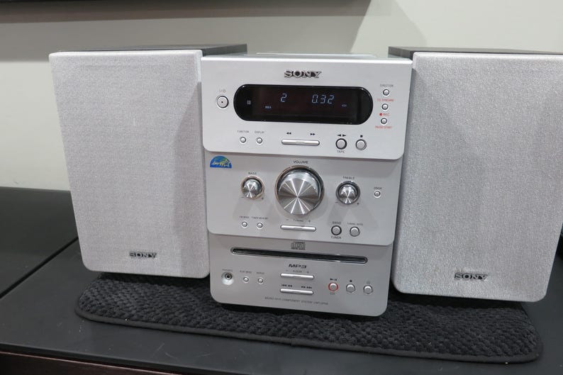 May include: A silver Sony stereo system with two speakers. The stereo has a digital display and various buttons and knobs. The speakers and stereo are on a black surface.