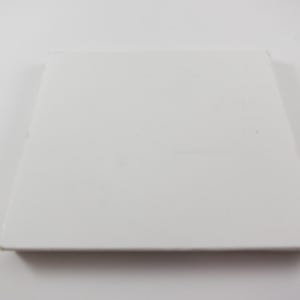 May include: A square, white ceramic tile with a slightly raised edge. The tile is plain and smooth, with a matte finish. The object is centered on a white background.