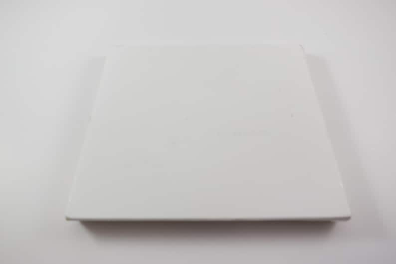May include: A square, white ceramic tile with a slightly raised edge. The tile is plain and smooth, with a matte finish. The object is centered on a white background.