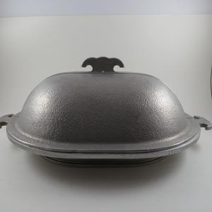 May include: A grey cast iron oval roaster with a textured exterior finish. The roaster features a domed lid with a central handle and two side handles on the base. The design is symmetrical with a heavy-duty construction and a matte metallic appearance.