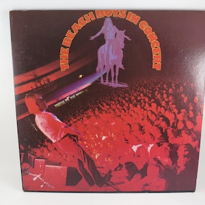 May include: Album cover for "The Beach Boys in Concert." The cover features a red and black color scheme with the band's name in orange. A silhouette of a person on a horse is above a crowd of people. A person is on stage in the foreground.