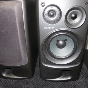 May include: Two black Sony bookshelf speakers. One speaker has a fabric grille cover with the Sony logo, while the other is exposed, revealing two small circular tweeters at the top and a larger woofer cone below, with the Sony logo printed on the front.