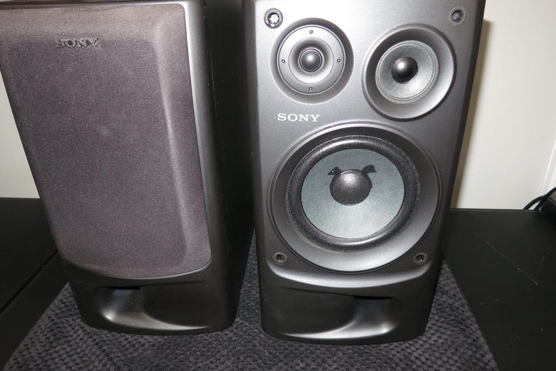 May include: Two black Sony bookshelf speakers. One speaker has a fabric grille cover with the Sony logo, while the other is exposed, revealing two small circular tweeters at the top and a larger woofer cone below, with the Sony logo printed on the front.