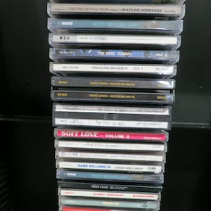 May include: A stack of compact discs with various album titles and artist names visible. The CDs are in clear plastic cases and include titles like "The Who Tommy," "Traveling Wilburys Vol. 1," and "Soft Love - Volume 3."