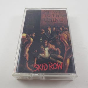 May include: A cassette tape with a black and red design. The front of the tape features a band of people in a dark setting. The text "Slave to the Grind" is printed in red on a white background. The band's name, "Skid Row", is printed in red at the bottom of the tape.