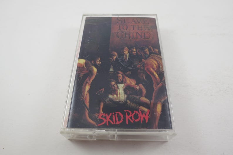 May include: A cassette tape with a black and red design. The front of the tape features a band of people in a dark setting. The text "Slave to the Grind" is printed in red on a white background. The band's name, "Skid Row", is printed in red at the bottom of the tape.
