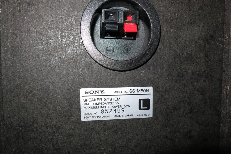 May include: The back panel of a black Sony SS-M50N speaker system featuring a circular terminal cup with one black and one red spring-clip connector. A white label displays technical specifications including 6 ohm impedance and 60W power.