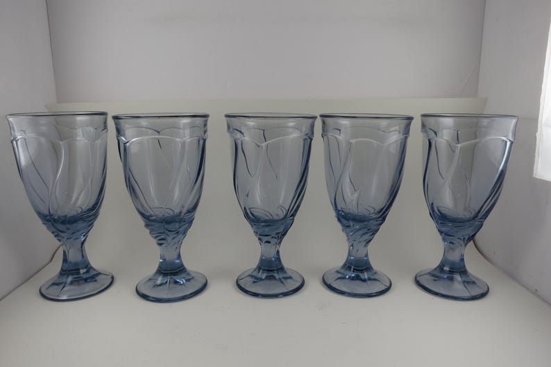 Pu&ograve; includere: Cinque calici d'acqua bluastri con un design decorativo a spirale. I bicchieri hanno una coppa a forma di tulipano, uno stelo e una base circolare. I calici sono di vetro e sono disposti in fila.