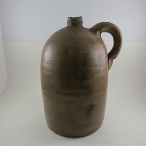 May include: A brown stoneware jug with a handle. The jug has a rounded body and a narrow neck with a slightly damaged rim. The handle is arched and attached to the side of the jug. The jug is approximately 14 inches tall.