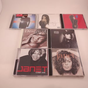 May include: A collection of eight compact discs featuring various artists, including Janet Jackson, Rhythm Nation, and Design of a Decade. The CDs are arranged in two rows, with the top row featuring four CDs and the bottom row featuring four CDs. The CDs are all in jewel cases and have various colors and designs.