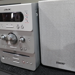 May include: Silver Sony stereo system with two speakers. The stereo has a digital display, buttons, and a CD player. The speakers and stereo are silver with the Sony logo visible. The stereo system is on a black surface.