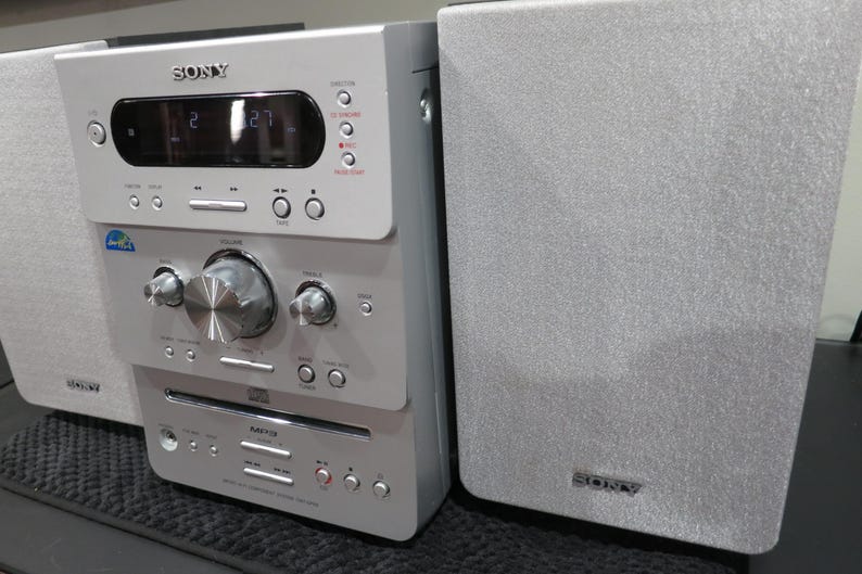 May include: Silver Sony stereo system with two speakers. The stereo has a digital display, buttons, and a CD player. The speakers and stereo are silver with the Sony logo visible. The stereo system is on a black surface.