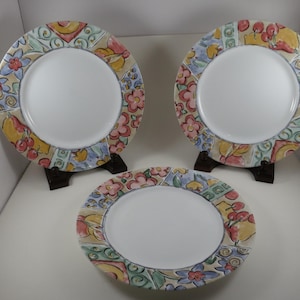 May include: Three round ceramic plates featuring a white center and a wide rim decorated with a colorful hand-painted pattern of red flowers, yellow fruit, and blue accents. Two plates are displayed on dark stands, while one lies flat in front.