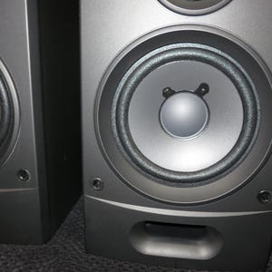 May include: Two dark grey speakers with circular speaker cones. The speakers have a simple, modern design with a rounded base and a small, rectangular port. They are likely for home audio or entertainment systems.