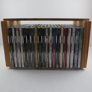 May include: Wooden CD storage rack filled with various music CDs. The rack is made of light-colored wood and has a handle on top. The CDs are in their jewel cases, with a variety of album covers visible.