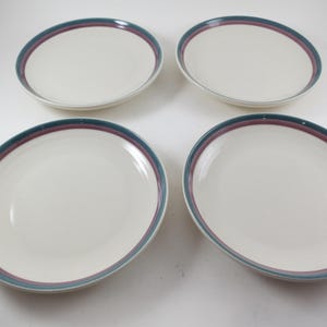 May include: Four off-white ceramic plates with a teal and burgundy border. The plates are oval-shaped and arranged on a white surface. The plates are clean and appear to be unused.