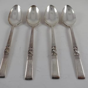May include: Four silver spoons with a decorative design on the handles. The spoons have a classic oval bowl shape and a detailed pattern near the base of the handle. The spoons are arranged on a white surface.