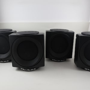 May include: Four black Wharfedale speakers. Each speaker is a cube shape with a circular speaker and a circular top. The speakers are sitting on a white surface.