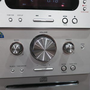 May include: Silver stereo system with multiple buttons and knobs for volume, bass, and treble control. Features a CD player and a digital display. Includes the text "FUNCTION", "DISPLAY", "TAPE", "REC", "PAUSE/START", "BASS", "VOLUME", "TREBLE", "TUNING", and "TUNER".