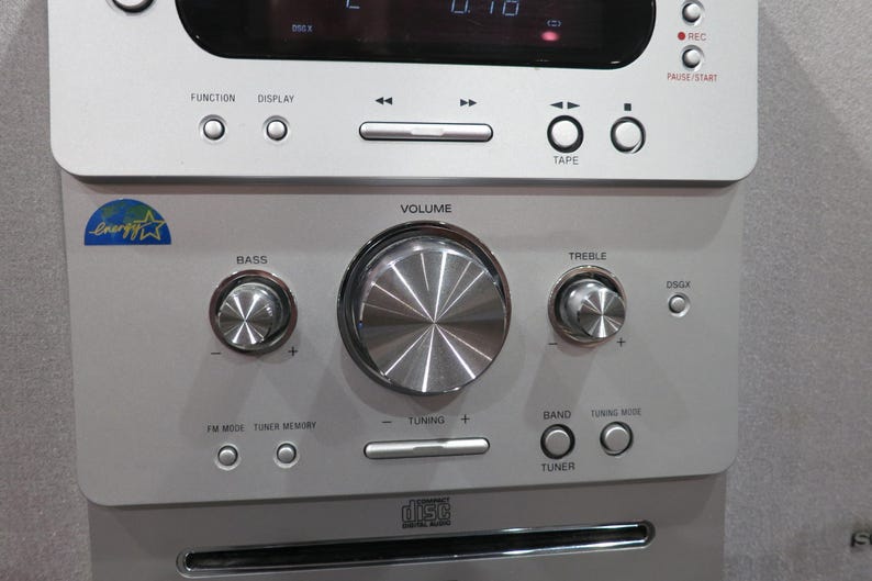 May include: Silver stereo system with multiple buttons and knobs for volume, bass, and treble control. Features a CD player and a digital display. Includes the text "FUNCTION", "DISPLAY", "TAPE", "REC", "PAUSE/START", "BASS", "VOLUME", "TREBLE", "TUNING", and "TUNER".